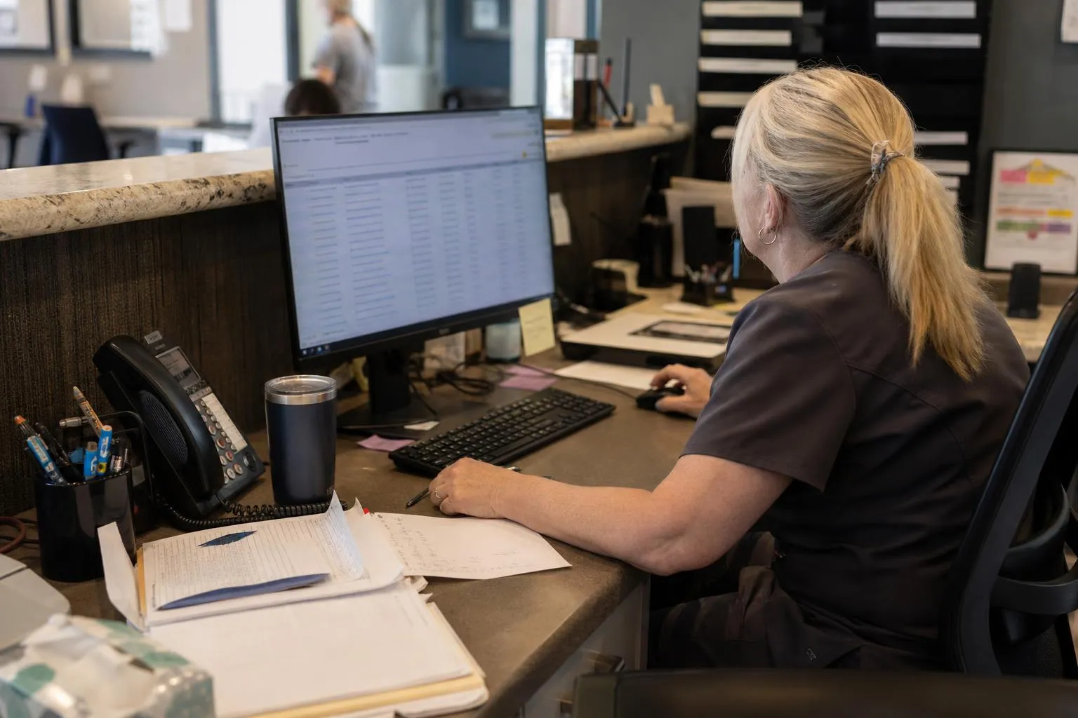 A practice manager reviews an overdue-patient spreadsheet on her monitor at the front desk
