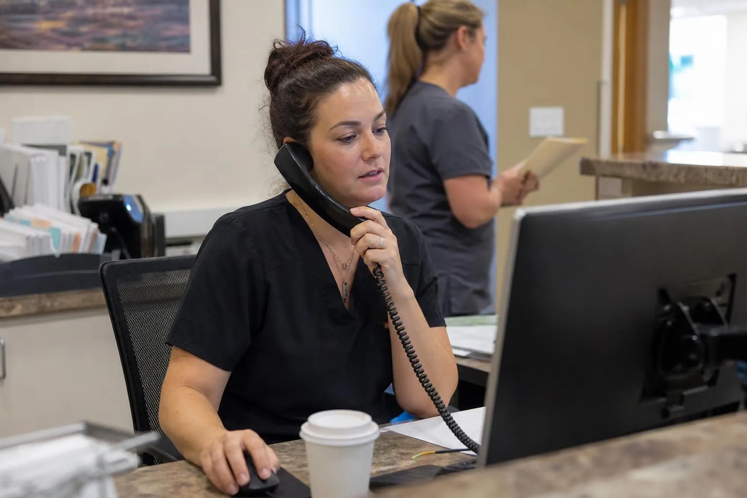 A front desk coordinator at a computer, checking appointment details during a patient phone call.