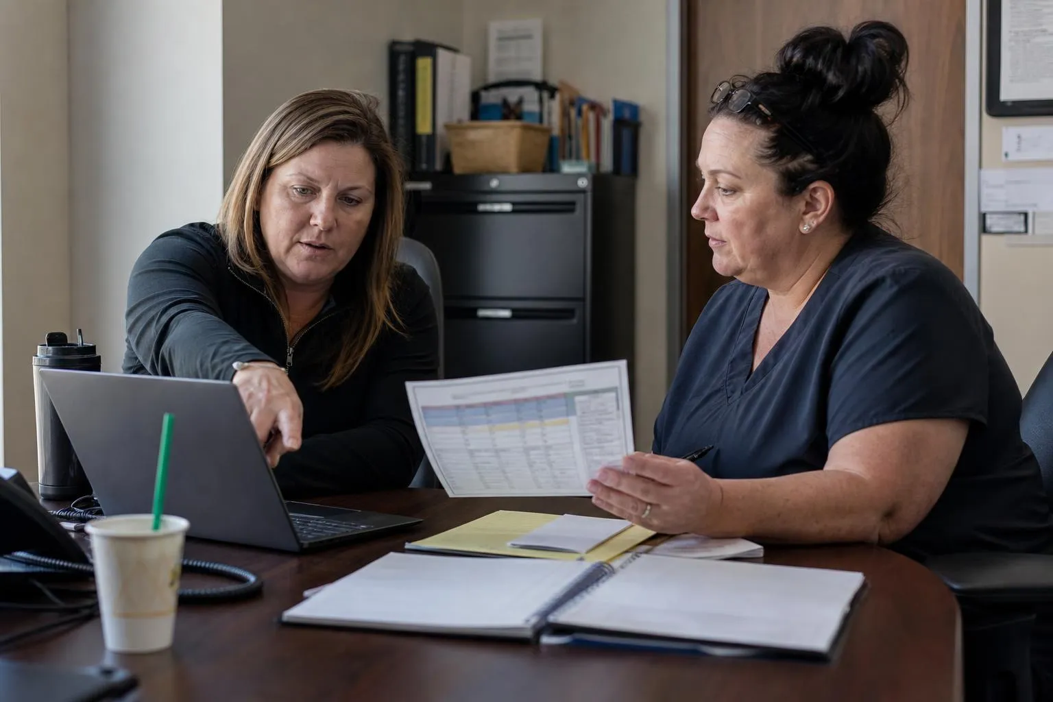 Two practice administrators comparing call coverage options on a laptop and printed notes at a conference table