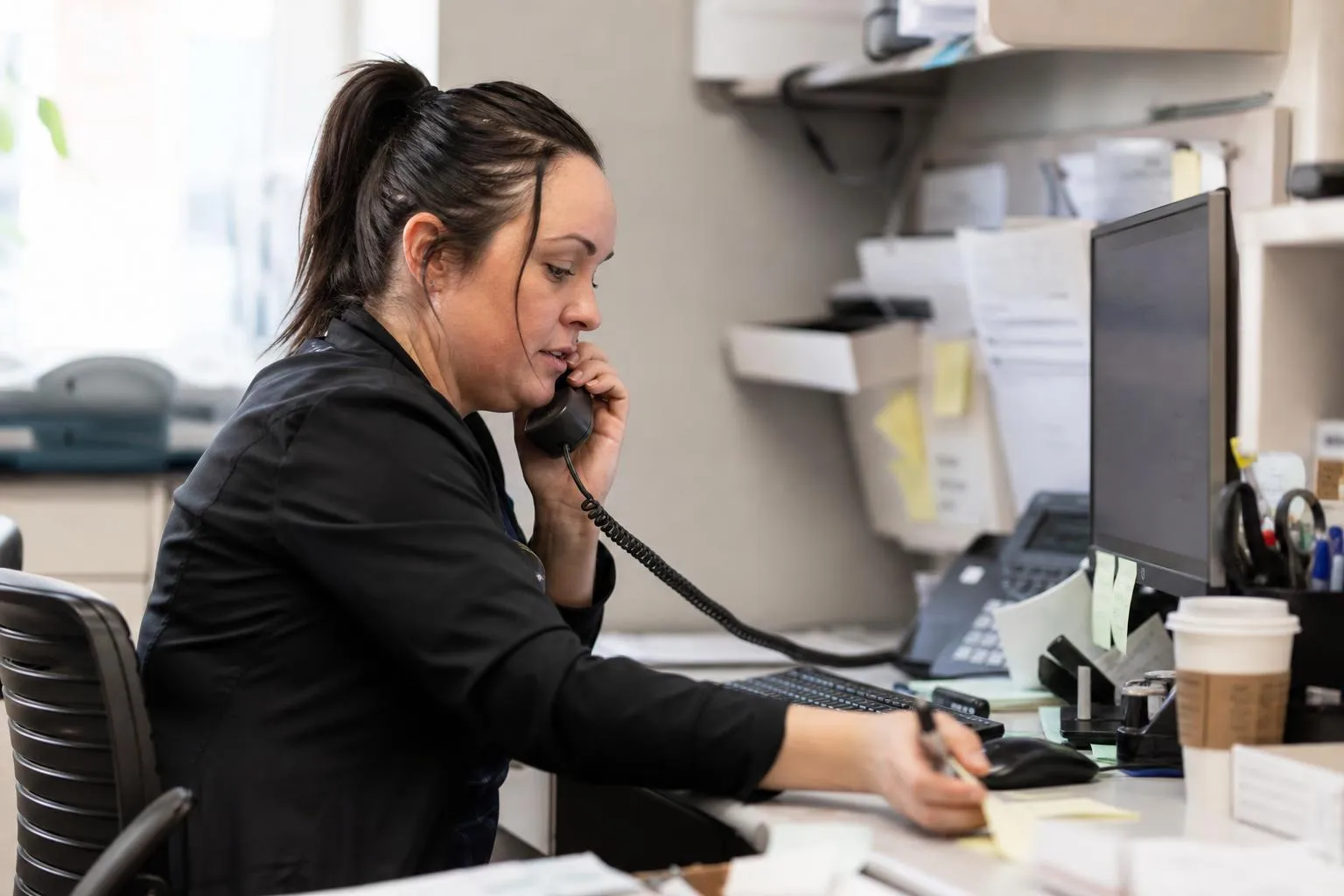 A front desk coordinator at a multi-provider medical practice answering a ringing phone while a scheduling screen sits open beside her