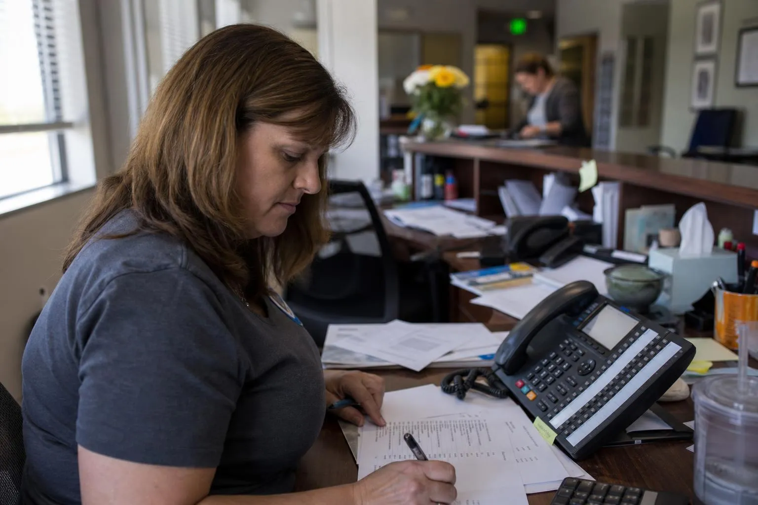 An office manager at a medical practice reviewing a callback list at her desk phone in the late afternoon