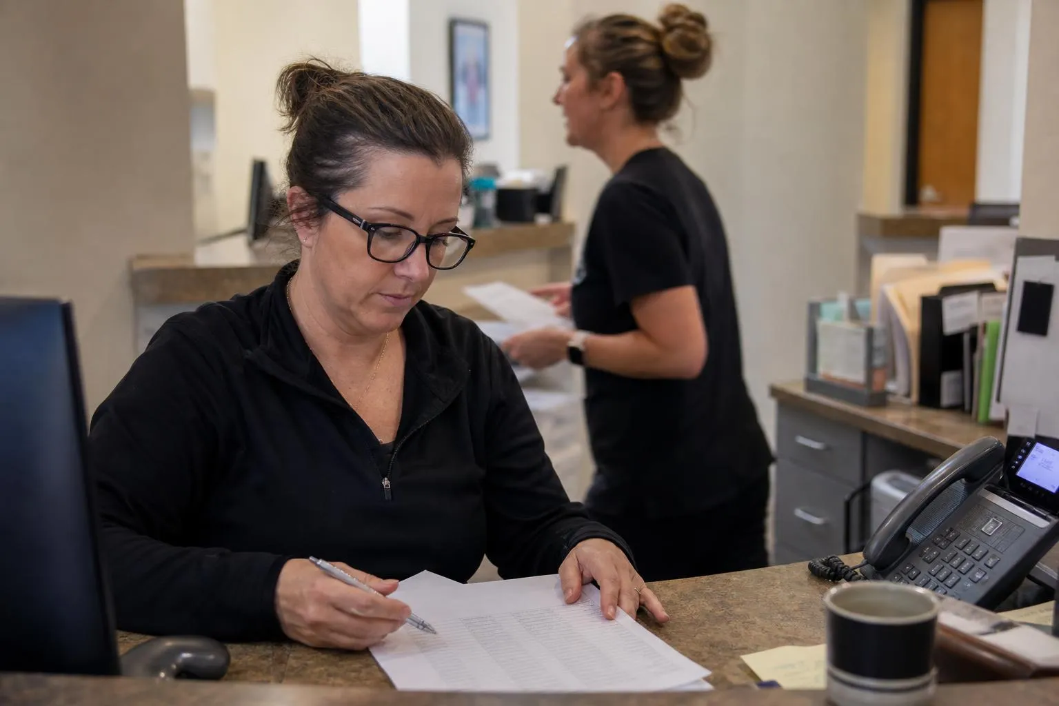 A practice administrator reviewing a printed overdue-patient report at a dental front desk while the phone lights up