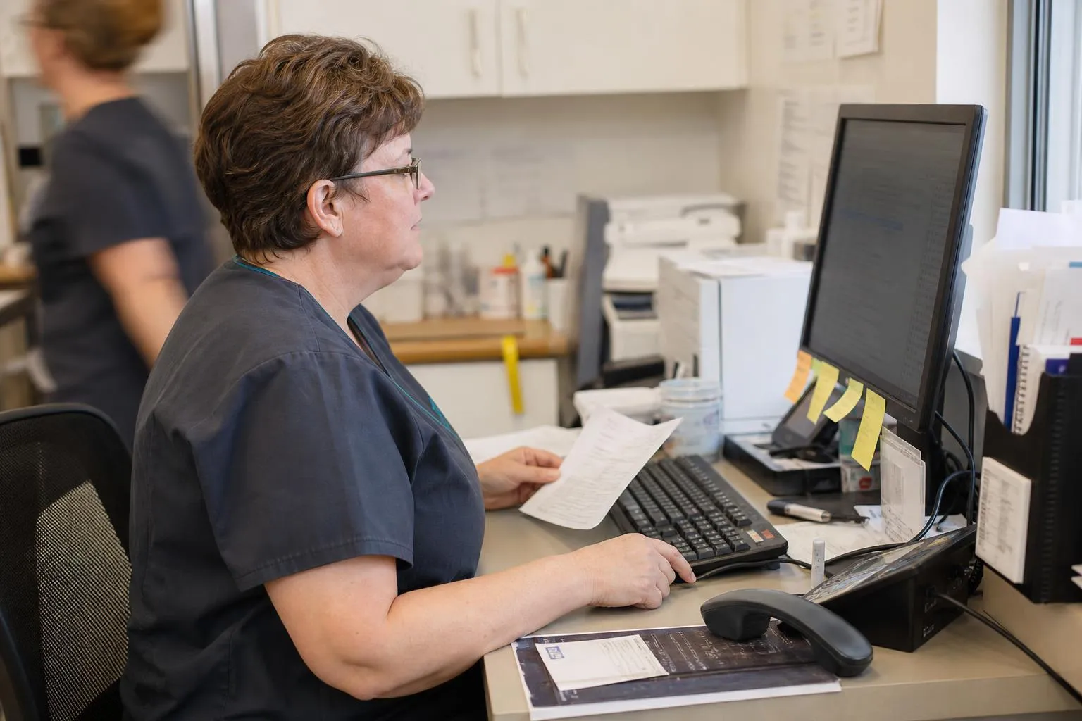 A referral coordinator at a specialty clinic working through a queue of new referrals on a desktop screen with a desk phone beside her