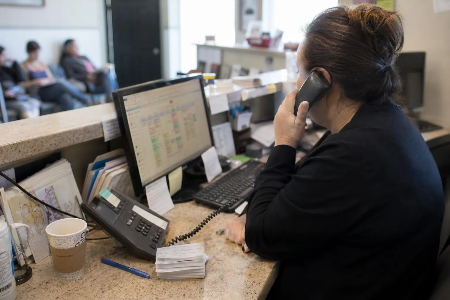 A front desk coordinator at a busy clinic answering the phone while viewing a scheduling calendar on screen