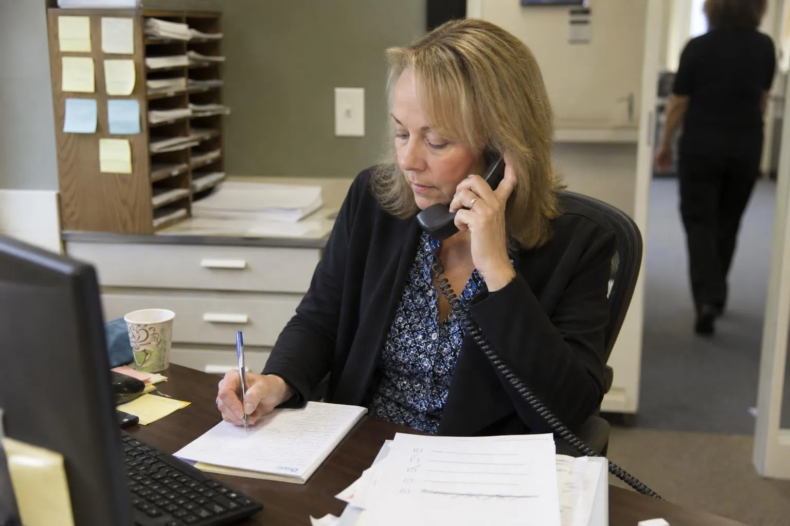An office manager on a phone call with a vendor, a printed checklist and handwritten notes on her desk
